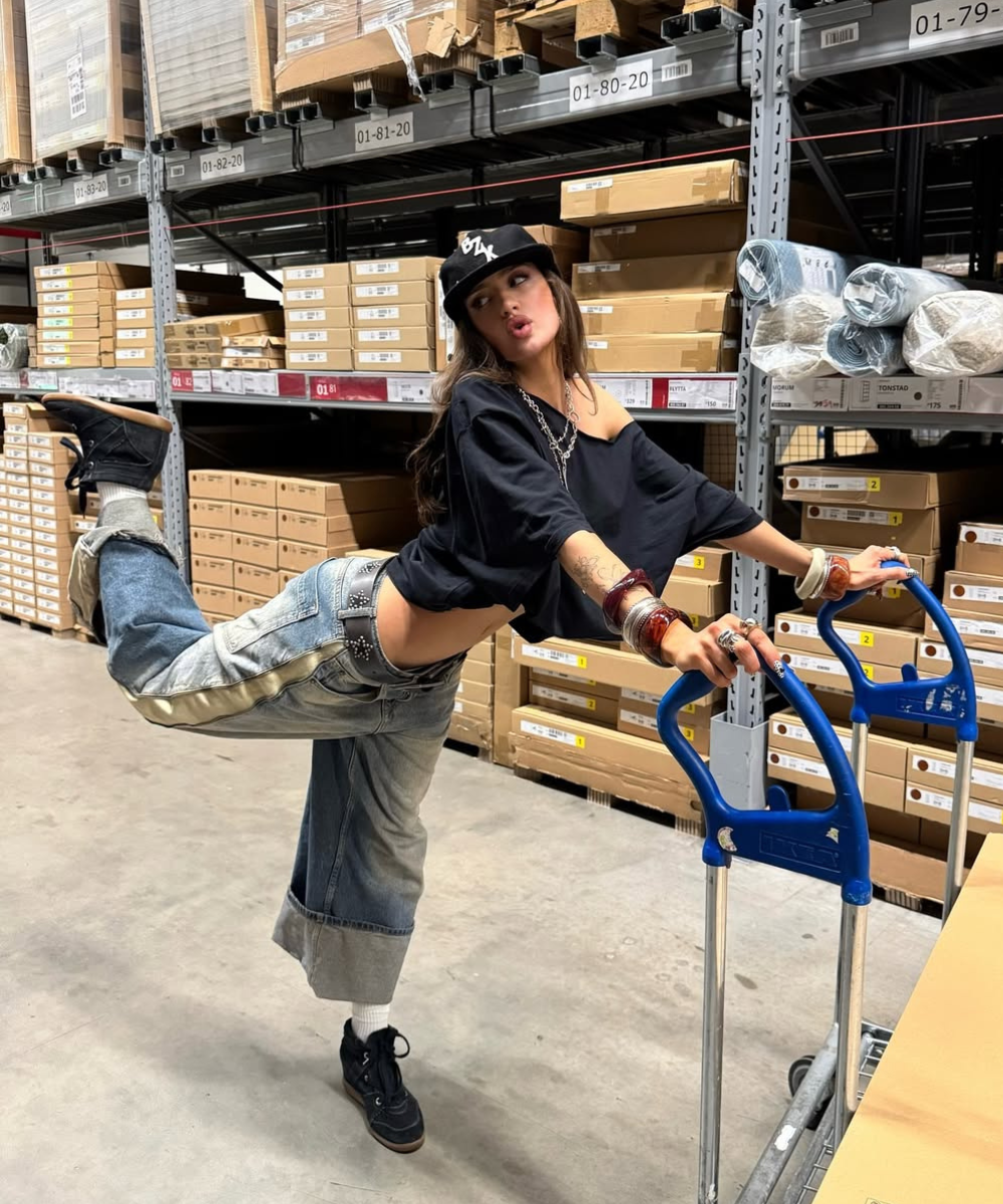 Pessoa em galpão industrial, vestindo jeans largos e camisa preta, equilibrada em carrinho de carga.