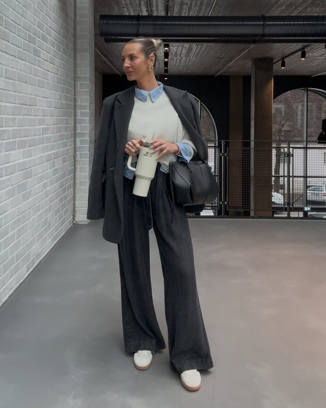 Mulher segurando o acessório funcional - garrafa térmica, vestida com casaco cinza e calça larga, em um ambiente moderno.
