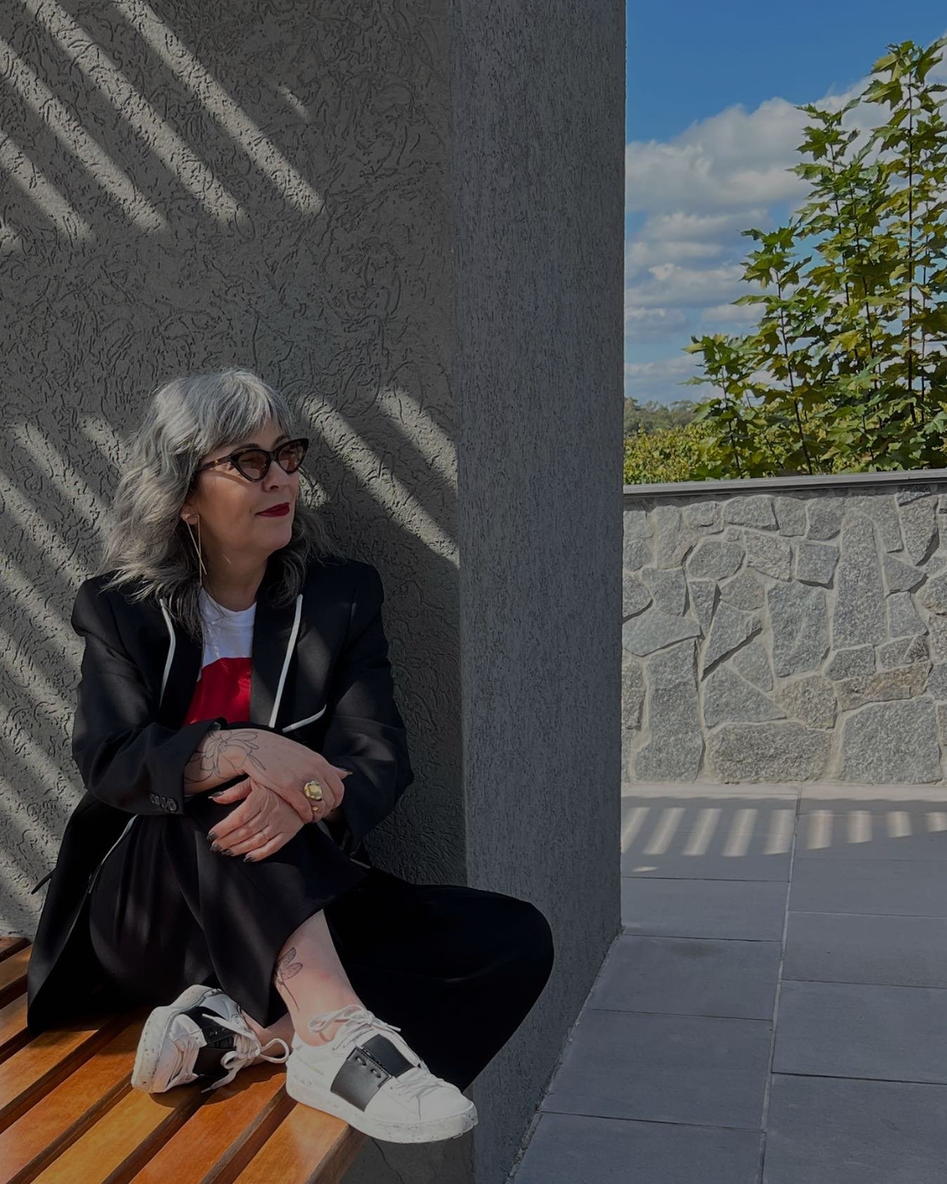 Mulher sentada em banco de madeira, cabelo grisalho em transição, usando blazer preto, sapato branco, céu azul ao fundo.