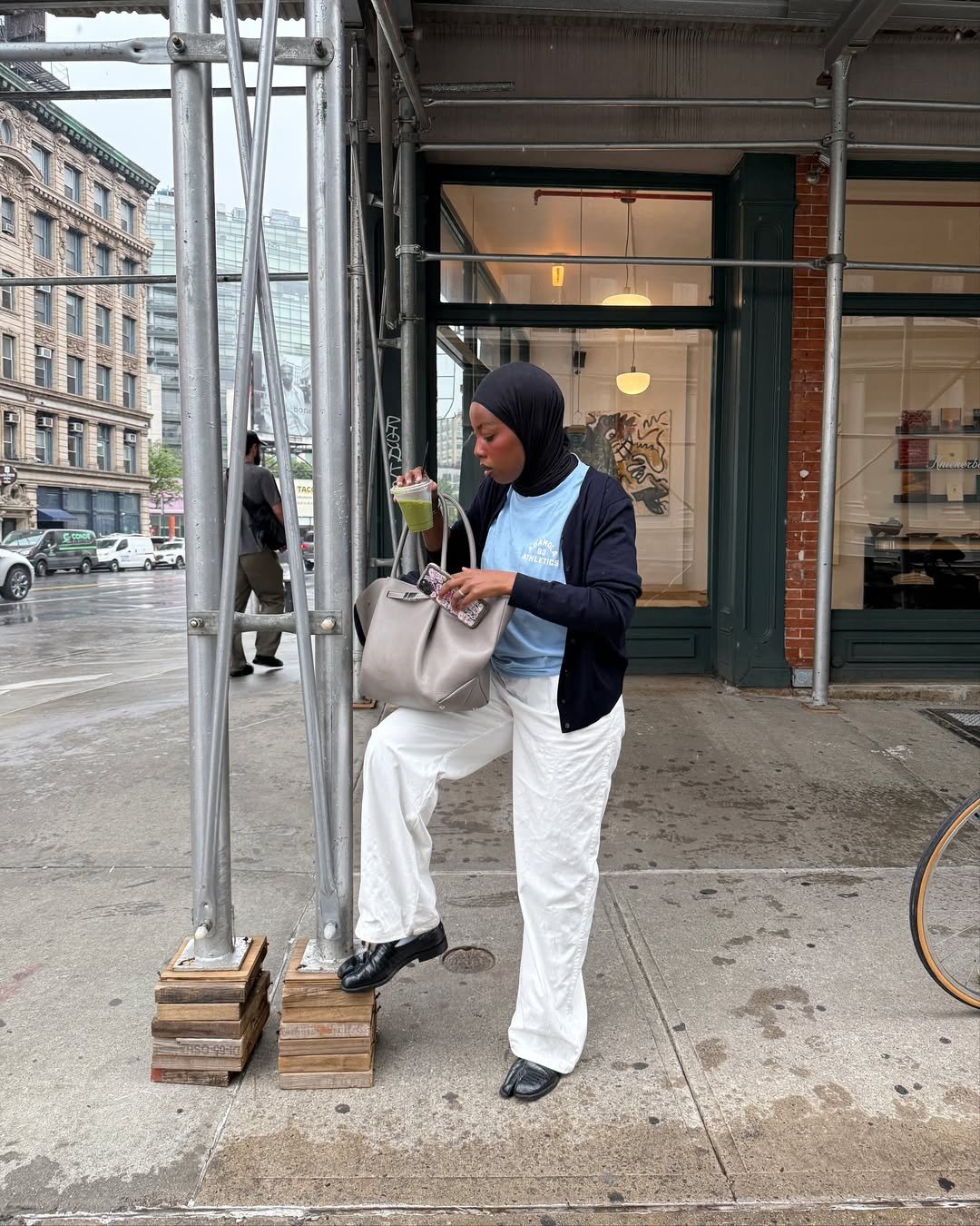 Pessoa de pé na calçada, usando um hijab, mexendo na bolsa com bebida na mão. Rua com estrutura metálica ao fundo.