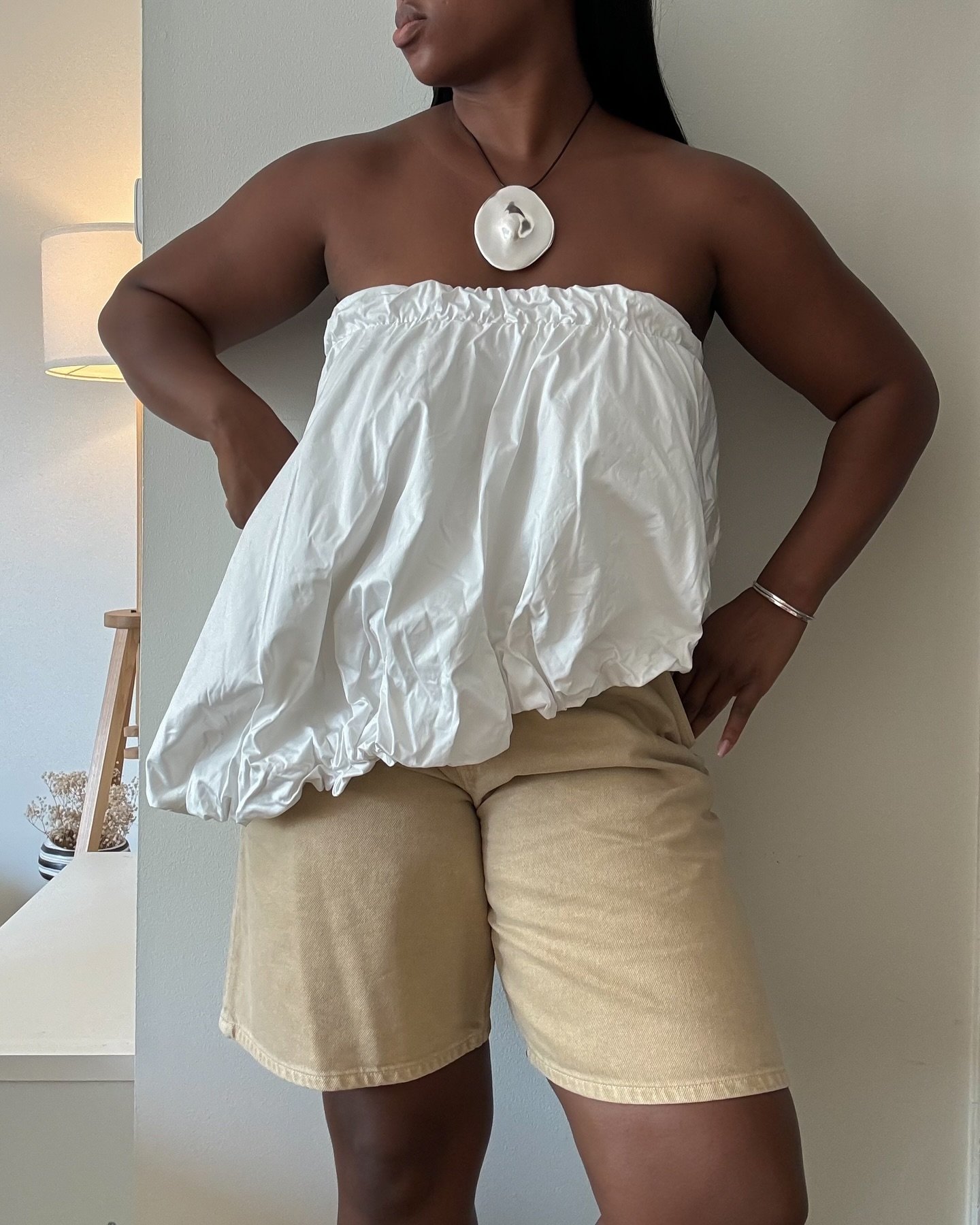 Mulher usando colar com pingente grande, vestido branco sem alças e bermuda bege, refletindo tendência de colar.