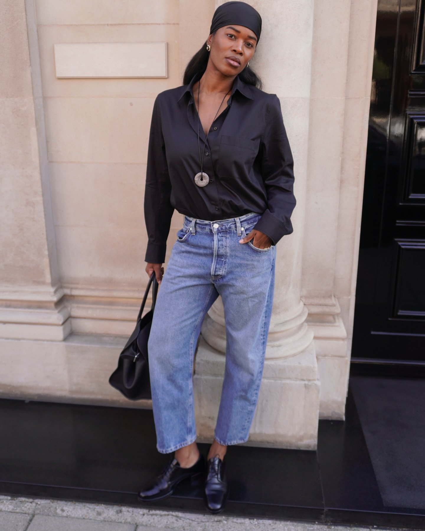 Mulher usando camisa preta, jeans e colar com pingente marcante, exemplificando tendência em acessórios.