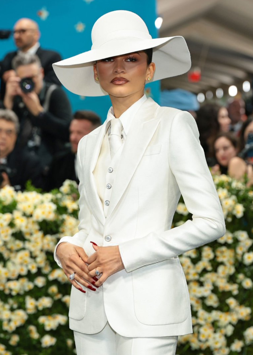 Pessoa em traje branco elegante com chapéu largo no Met Gala 2025, cercada por flores e fotógrafos.