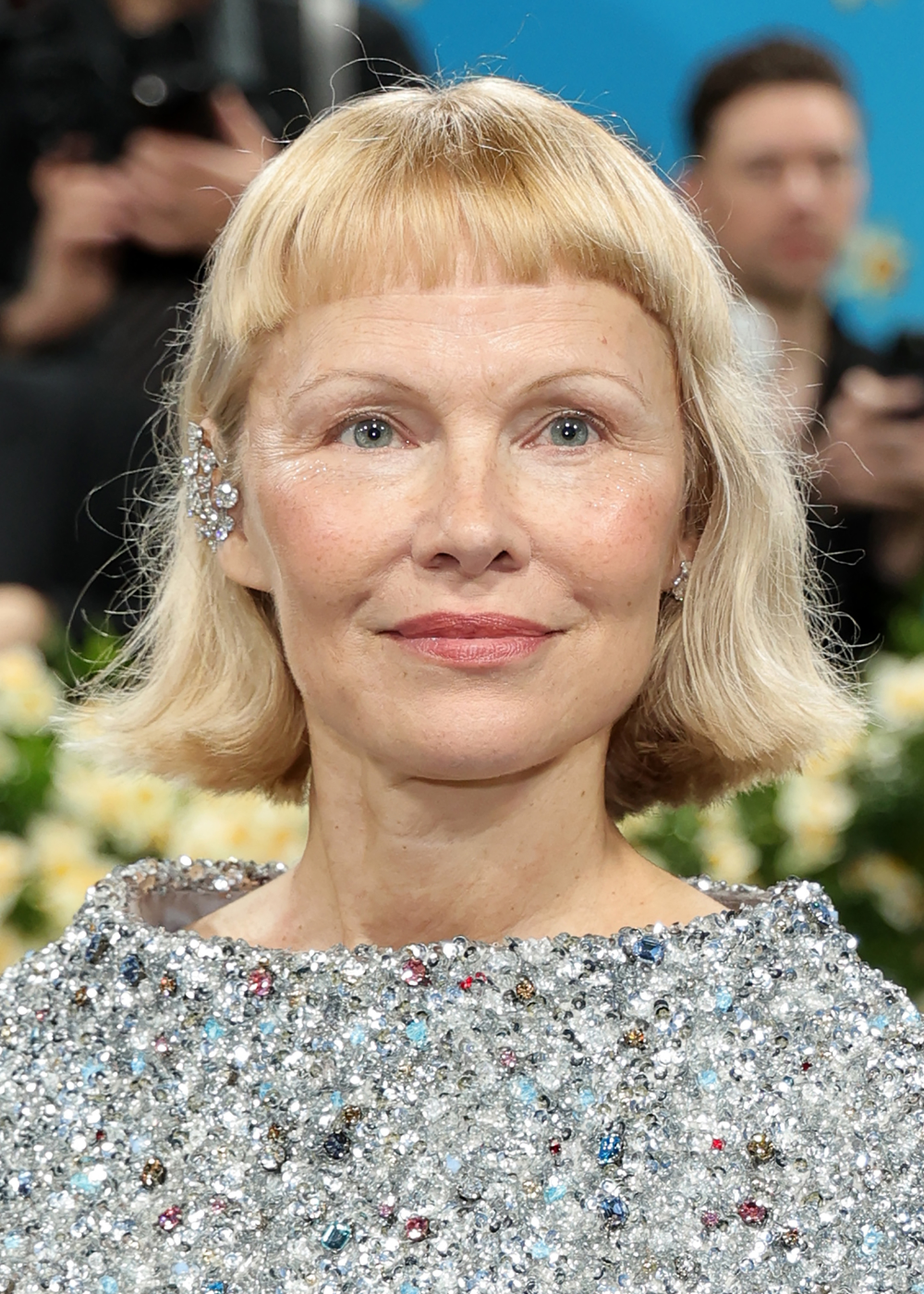 Pessoa em vestido brilhante no Met Gala 2025, com cabelos loiros curtos e acessórios elegantes. Fundo desfocado.