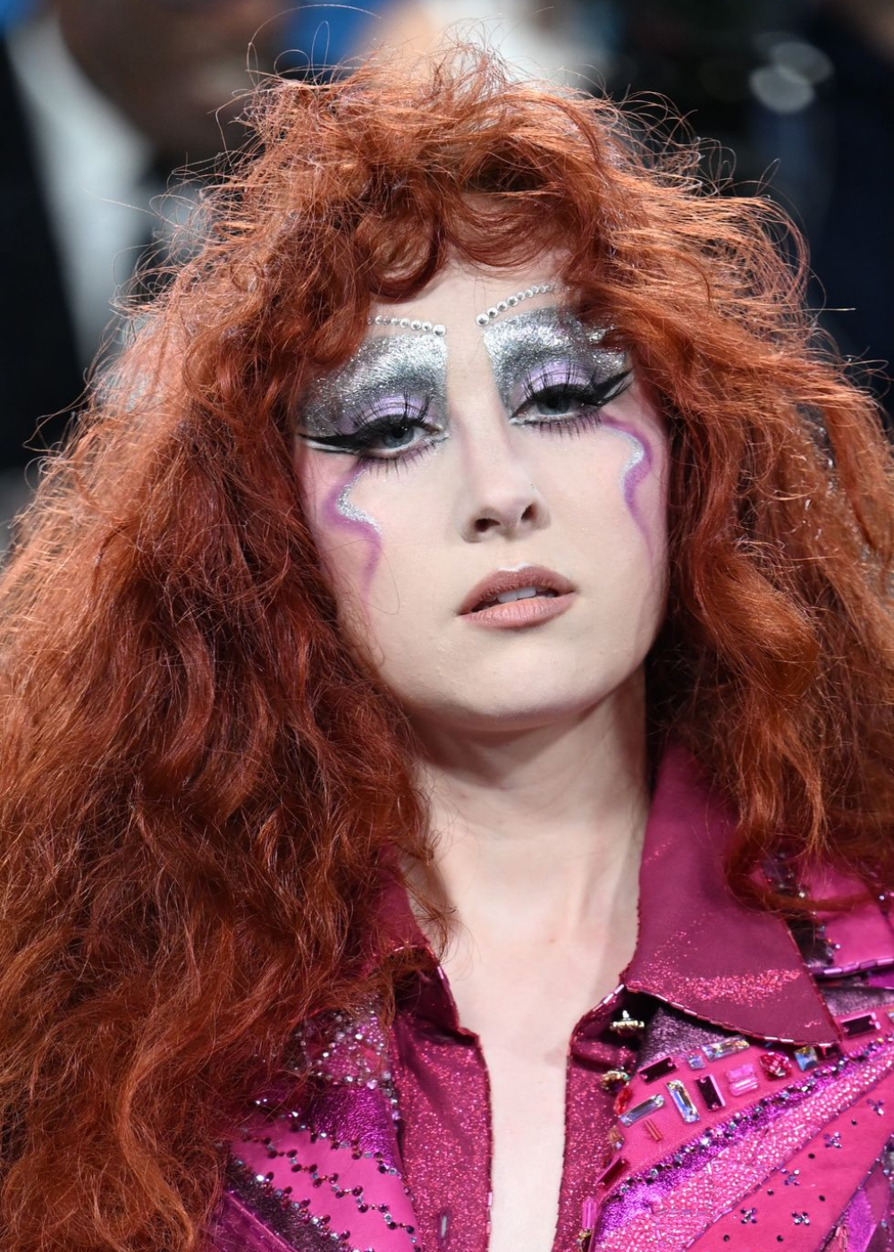 Pessoa com cabelo ruivo e maquiagem metálica elaborada usa roupa brilhante roxa no Met Gala 2025.