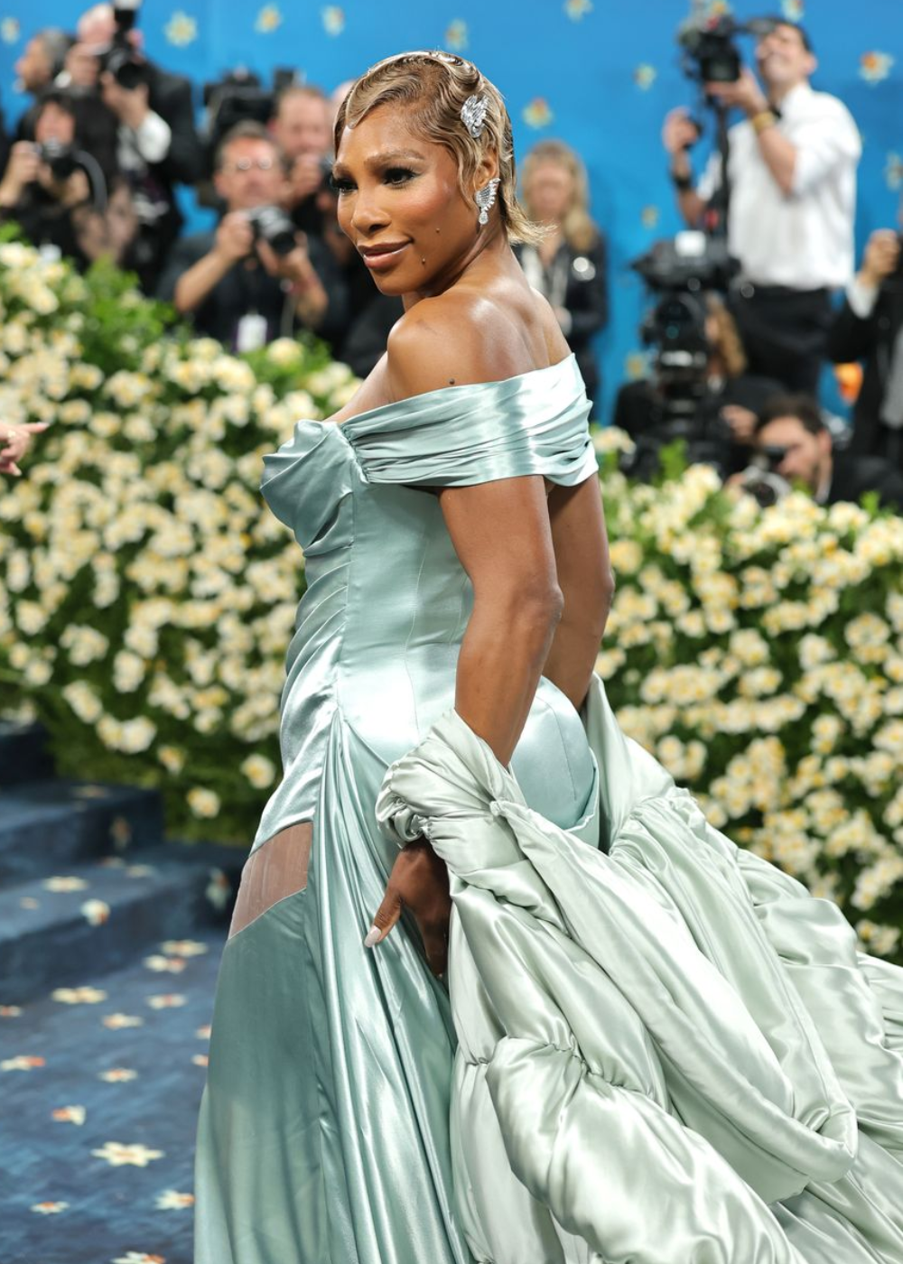 Vestido azul brilhante com detalhes prateados e volumosos em evento do Met Gala 2025, cercado por flores e fotógrafos.