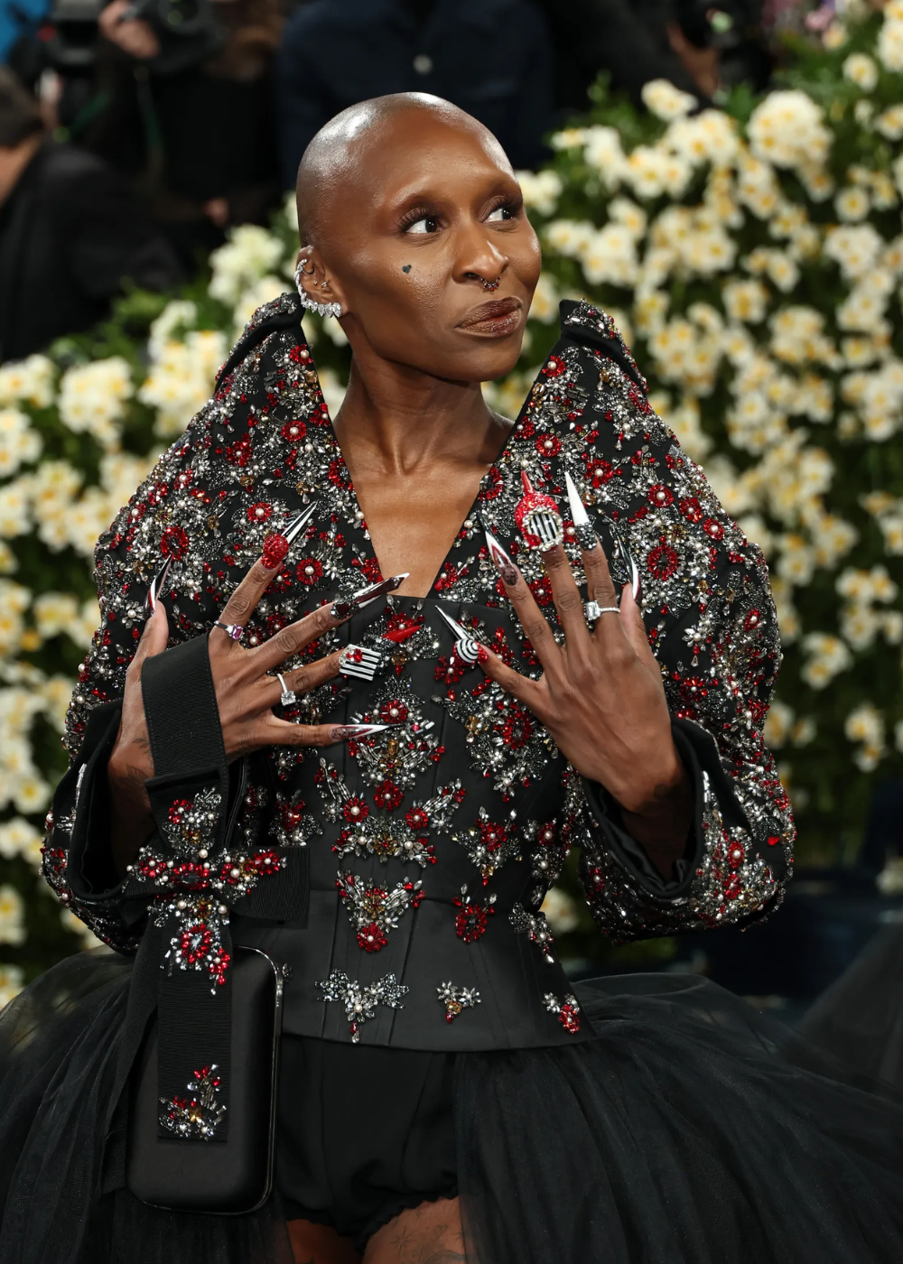 Vestido preto com pedraria brilhante e unhas decoradas em destaque no Met Gala 2025, fundo florido ao fundo.