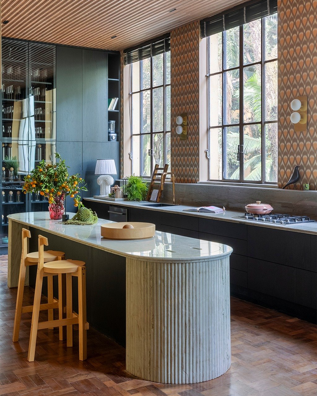 Cozinha moderna com design elegante. Ilha central com bancadas claras e banquetas. Grandes janelas iluminam o ambiente. CASACOR 2025.