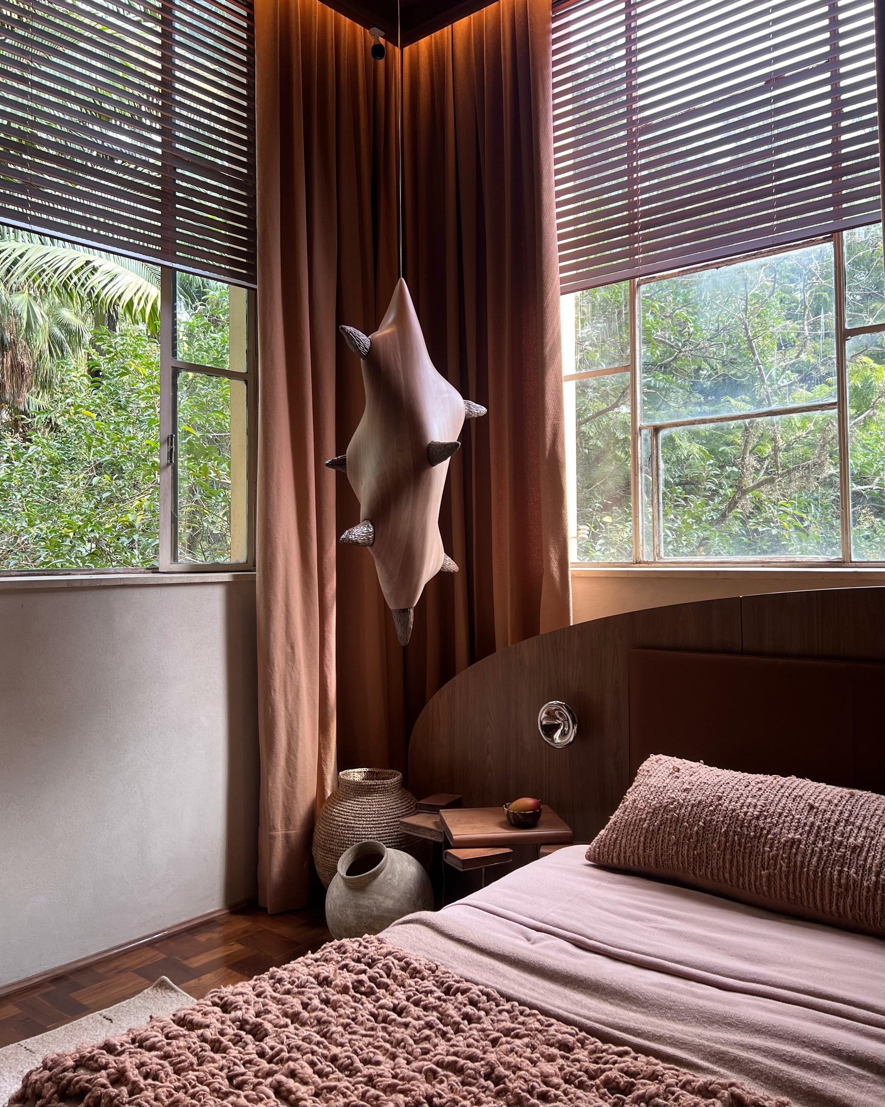 Quarto aconchegante com cama de tecido marrom, luminária escultural, cestos decorativos e grandes janelas. CASACOR 2025.