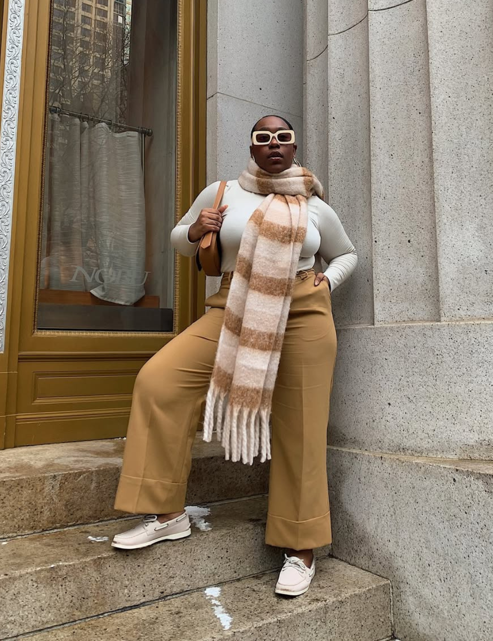 Pessoa com peças estilosas como óculos e cachecol, em look tendência, posa em escadas diante de prédio elegante.