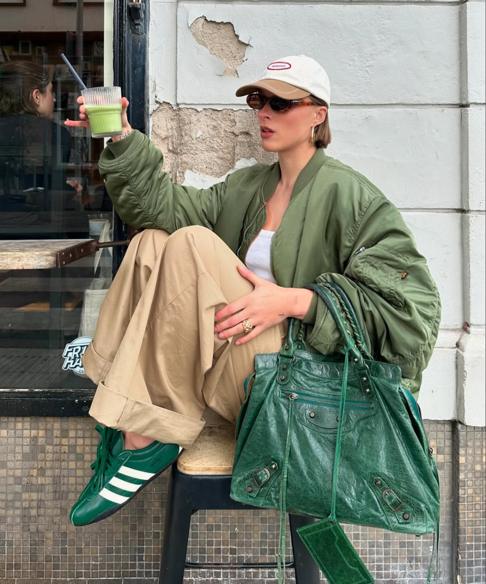 Pessoa sentada com bebê verde, calça bege e tênis femininos estilosos verdes, segura bebida. Bolsa verde ao lado.