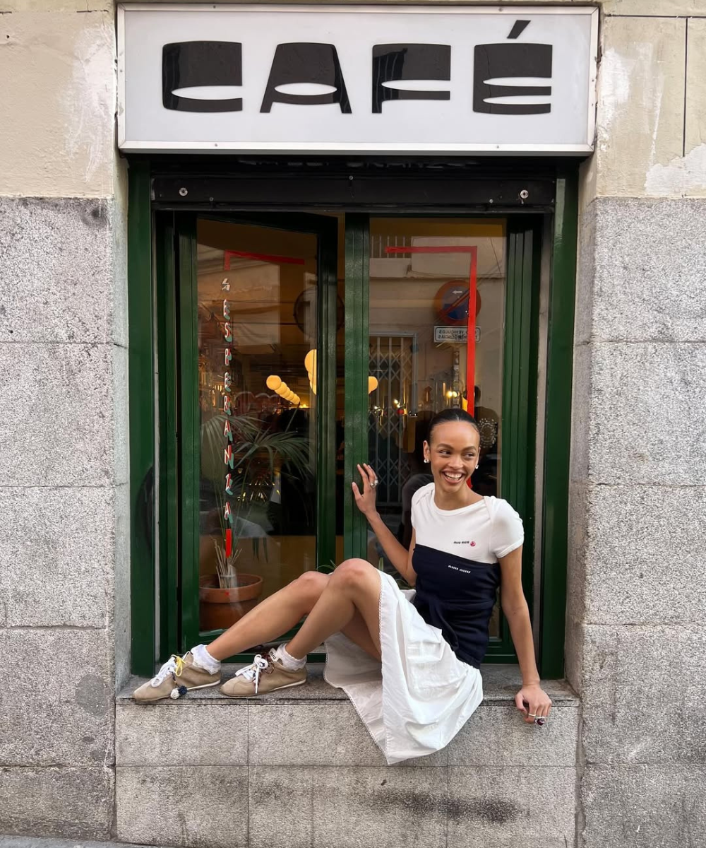 Pessoa sorrindo sentada em parapeito de janela de café, usando vestido e tênis femininos estilosos.