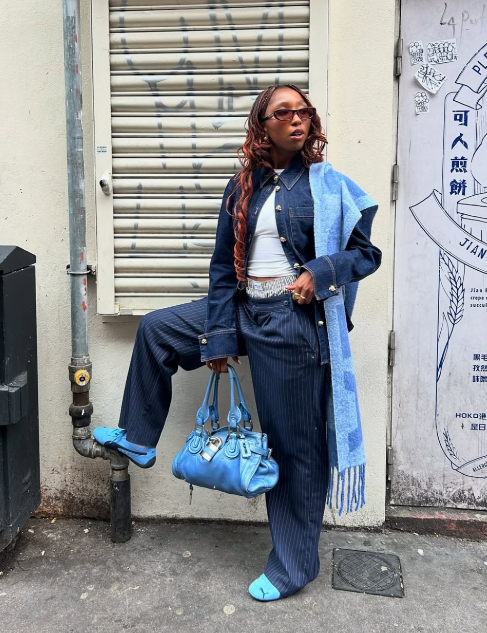 Pessoa usando conjunto jeans e lenço azul, com bolsa azul e tênis feminino colorido. Fundo de parede grafitada.
