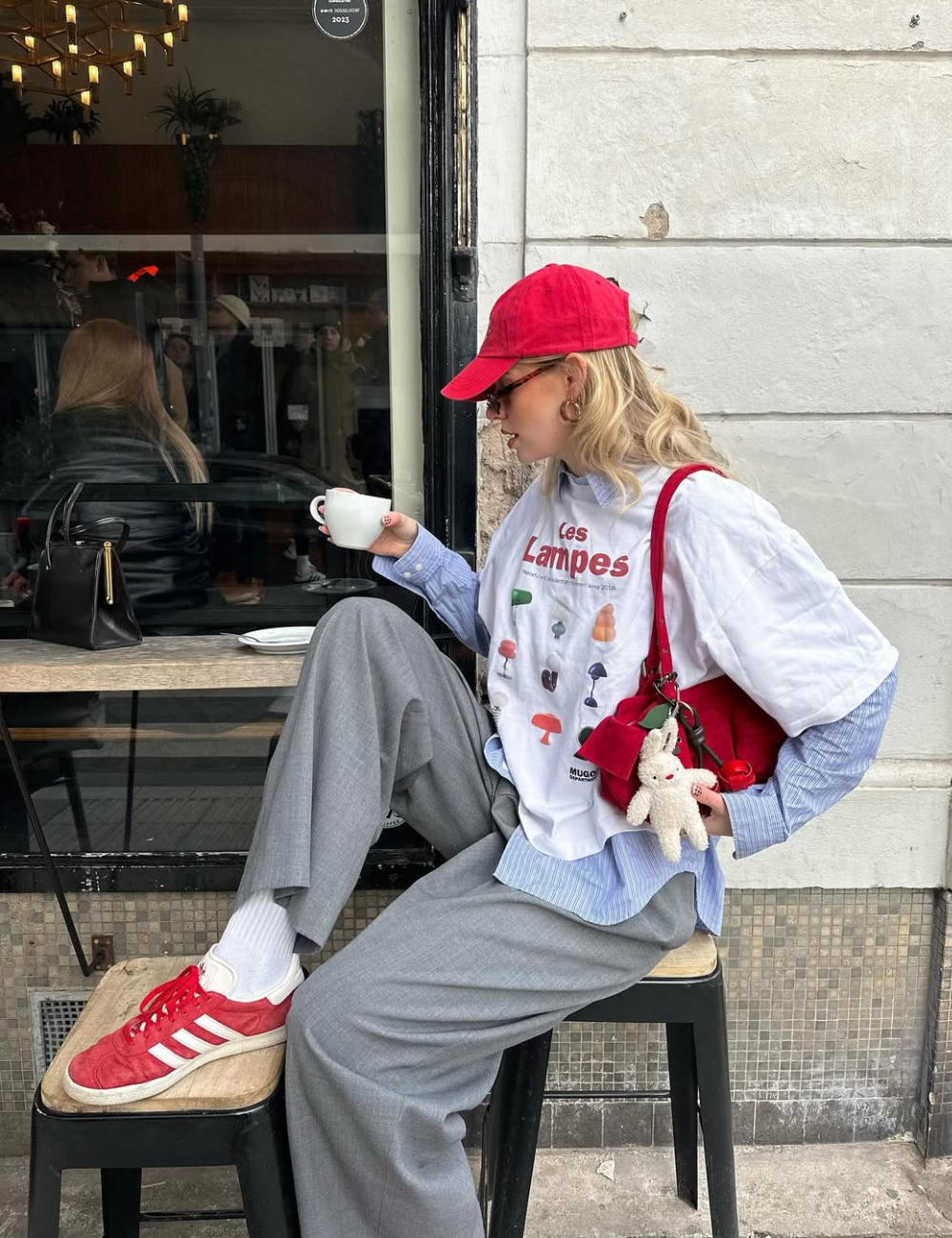 Pessoa sentada em café, usando boné vermelho e segurando xícara. Ela veste calças cinza e tênis vermelhos.