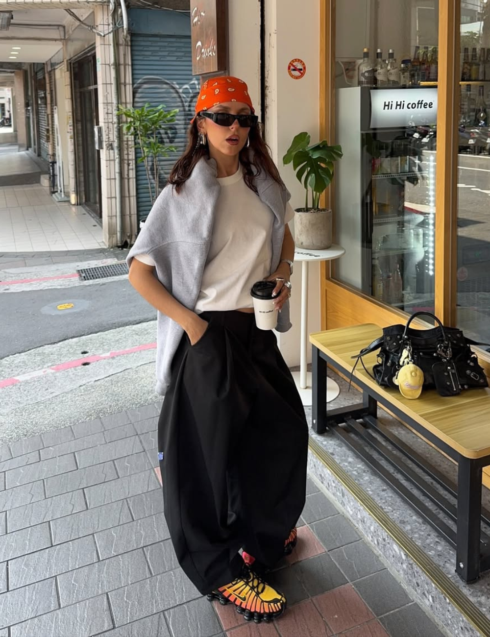 Mulher com bandana laranja, camiseta branca e calça preta, segura café. Usa tênis feminino colorido.
