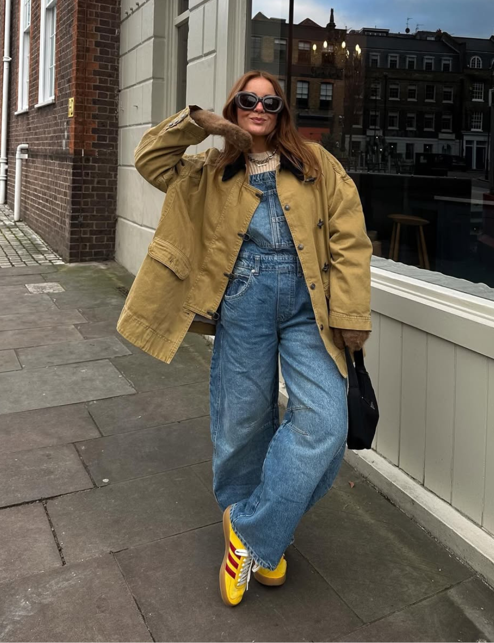 Mulher em macacão jeans, casaco bege, óculos escuros e tênis feminino colorido. Ela está sorrindo e posando na calçada.