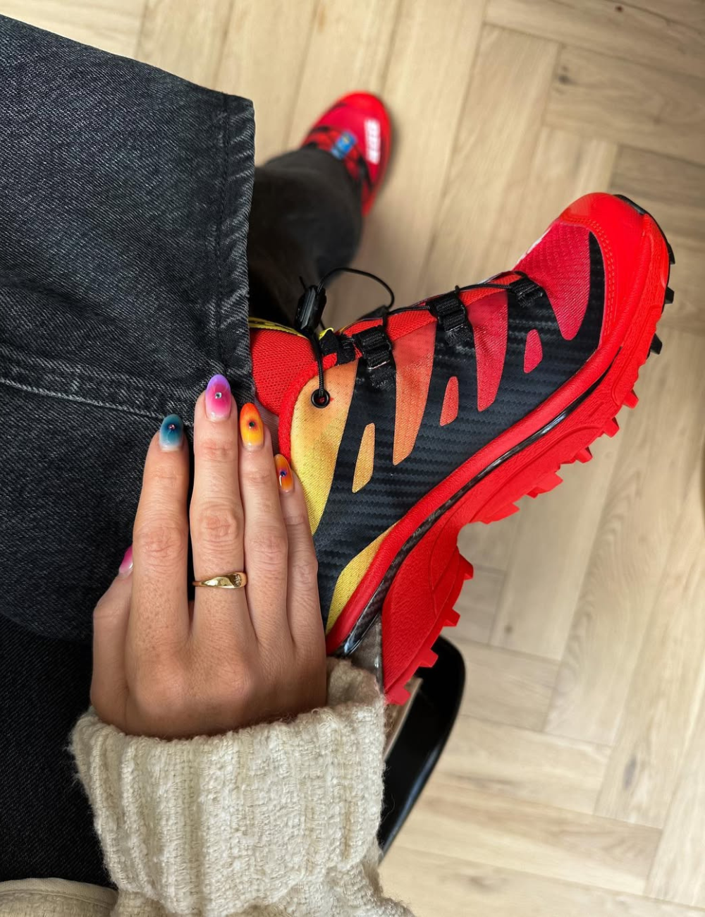 Tênis vermelho e preto com design ousado, acompanhados de unhas coloridas e anel dourado.