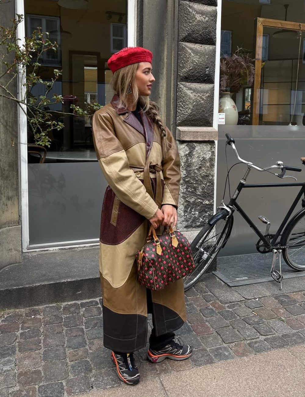 Mulher com look estiloso usa tênis polêmico tendência, casaco longo em tons de marrom e boina vermelha ao lado de bicicleta.