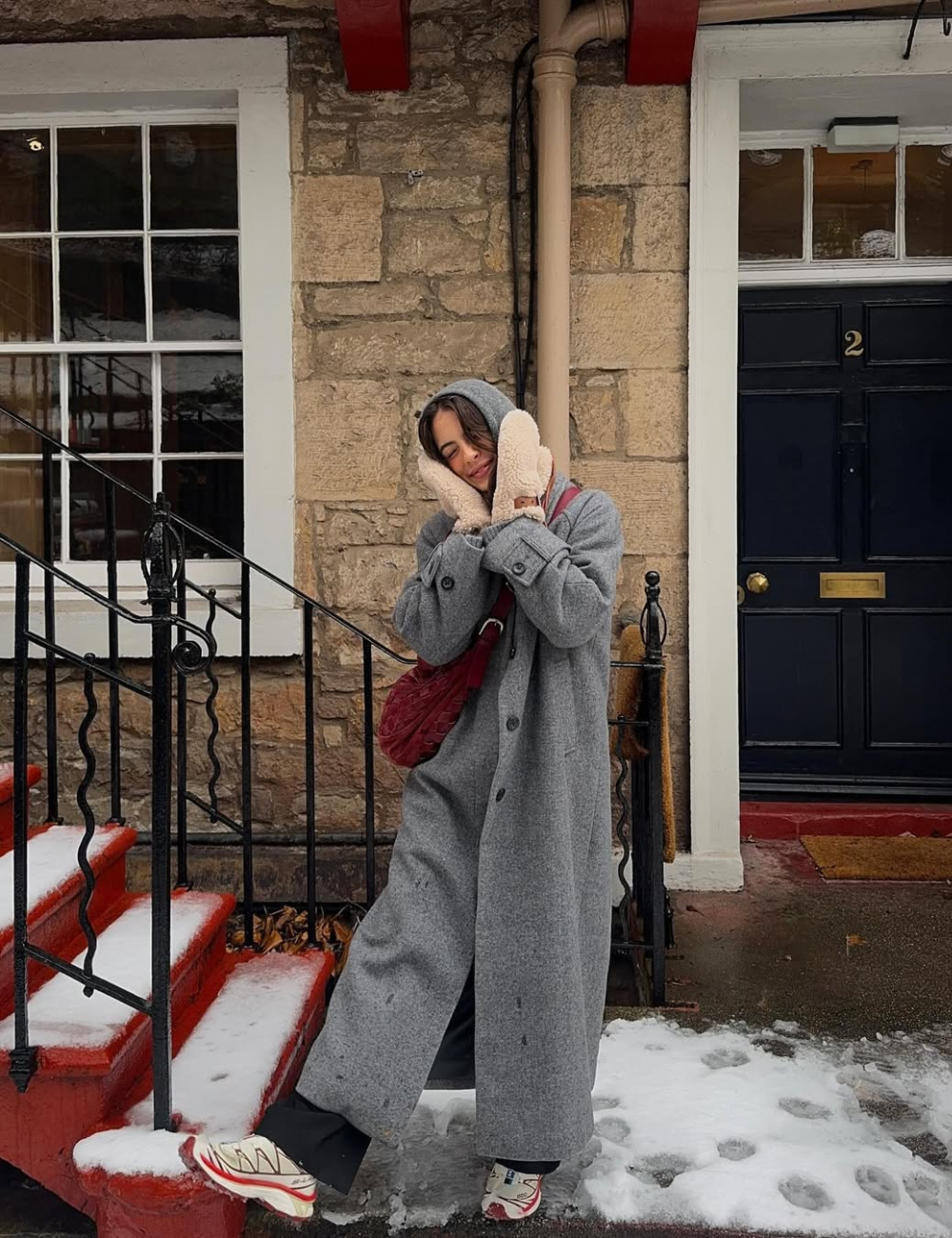 Pessoa com casaco cinza longo, luvas, bolsa vermelha e tênis estiloso em uma escada com neve. 