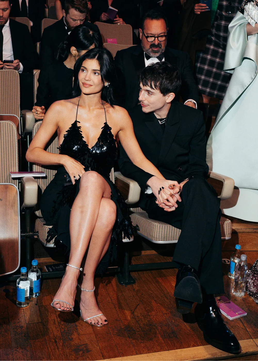 Kylie Jenner e Timothée Chalamet estão sentados lado a lado em um auditório, vestidos elegantemente. Eles parecem interagir de forma amigável.