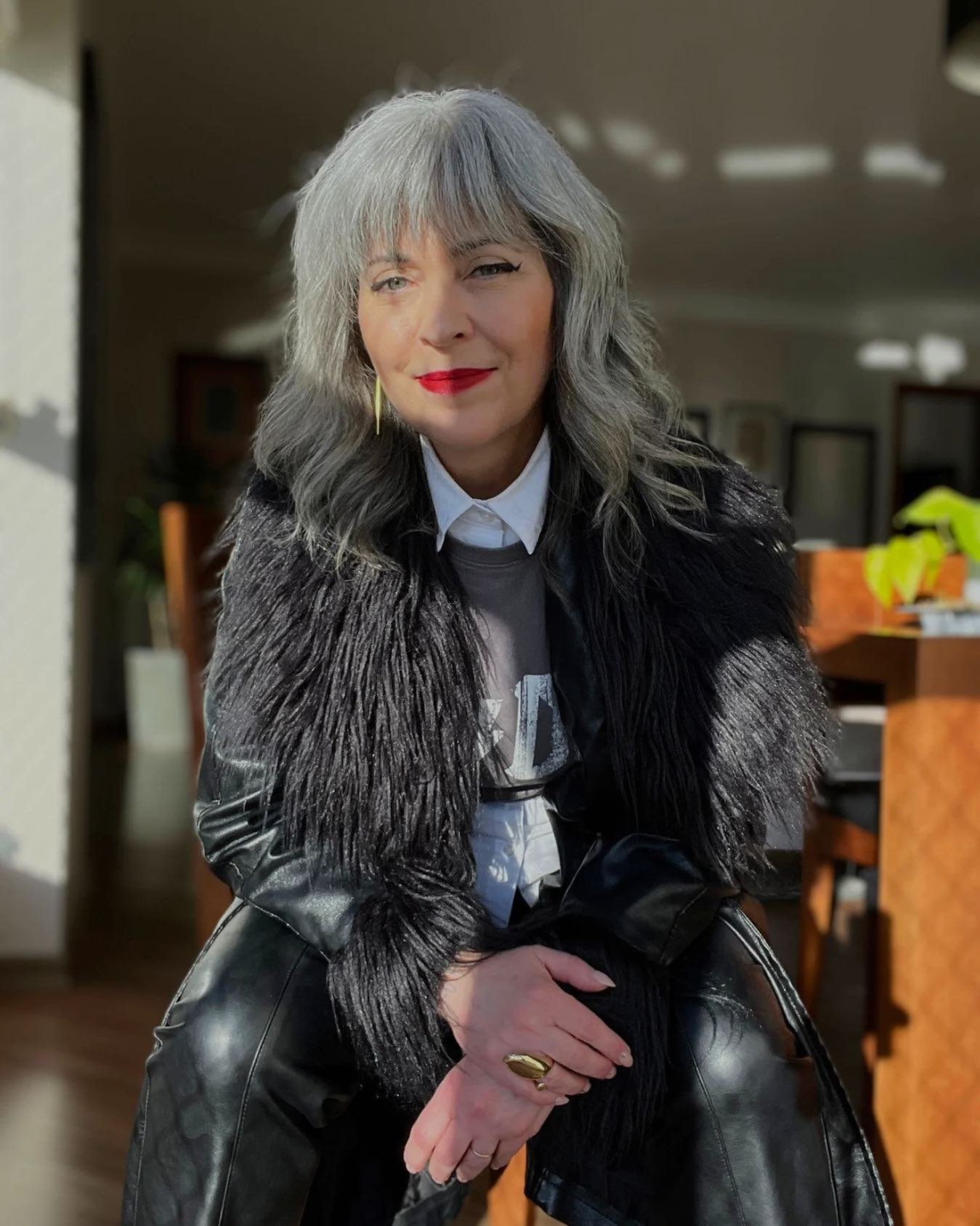 Mulher com cabelo grisalho, aparentando estar em transição, veste roupa preta em ambiente iluminado.