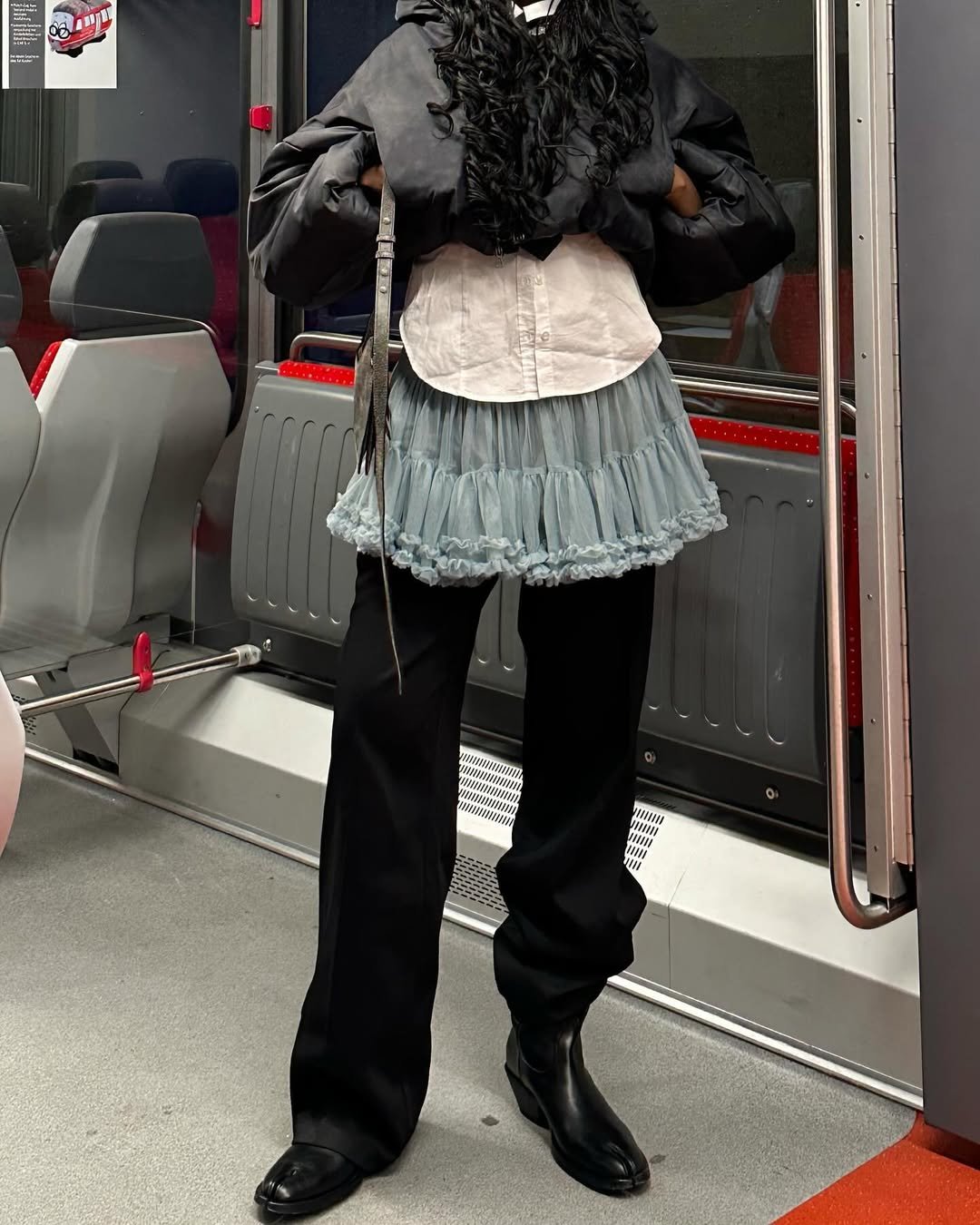Pessoa em transporte público usa saia rendada sobre calça preta, combinando camisa e jaqueta. Truque de styling fácil.