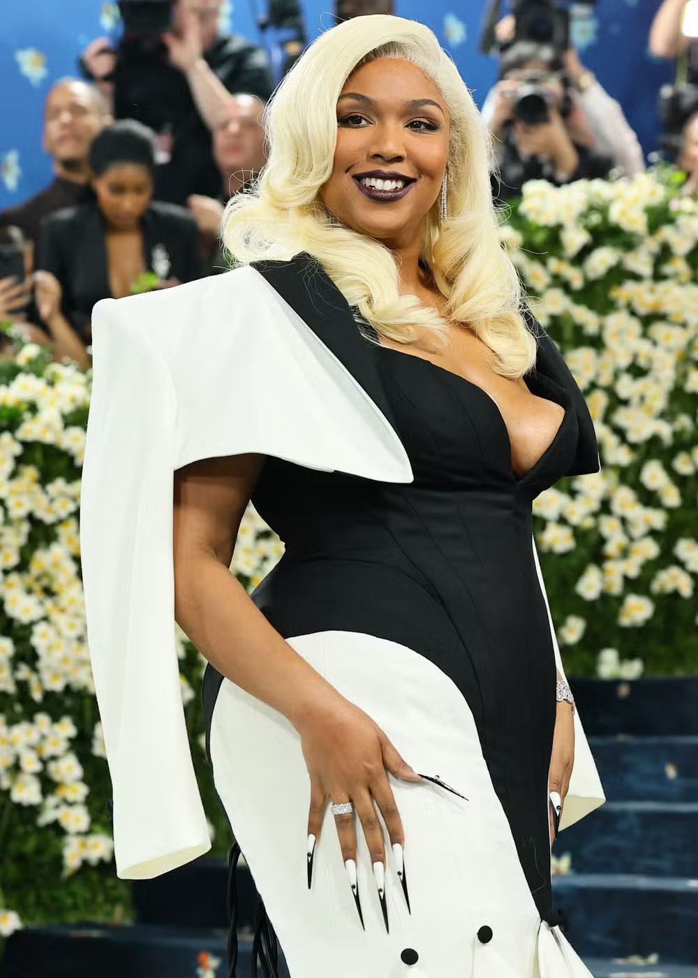 Mulher sorrindo em evento, usando vestido preto e branco. Suas unhas maximalistas são longas e chamativas.