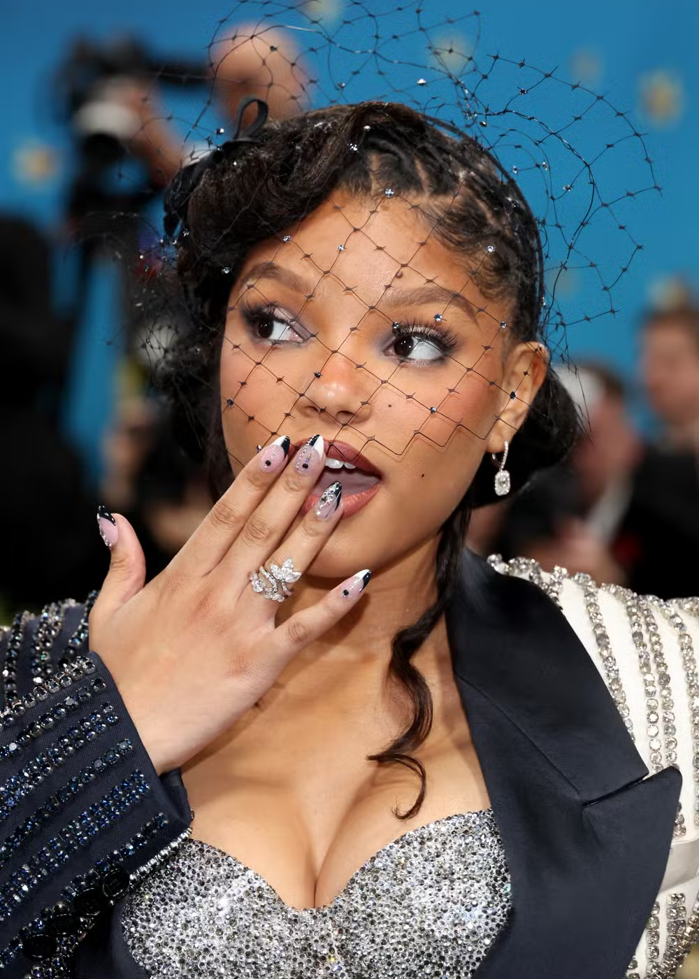 Jovem com véu e unhas maximalistas decoradas; ela usa anel chamativo, vestido com brilhos e expressão surpresa.