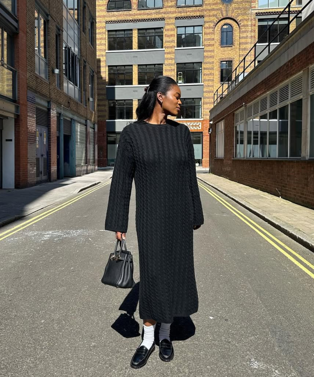 Mulher de vestido chique preto em pé na rua, segurando uma bolsa. Edifícios ao fundo.