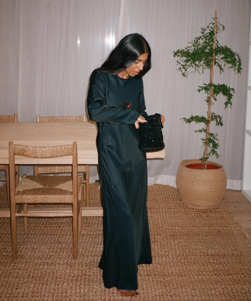 Mulher usando vestido chique preto, segurando uma bolsa, em sala decorada com cadeira de madeira e planta.