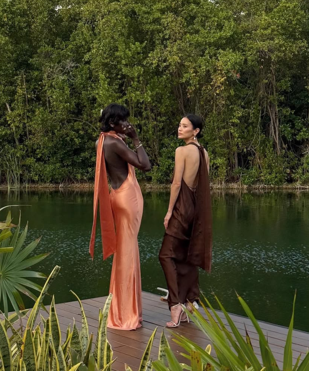 Duas mulheres em vestidos formais longos, um laranja e outro marrom, de frente para um lago rodeado por vegetação.