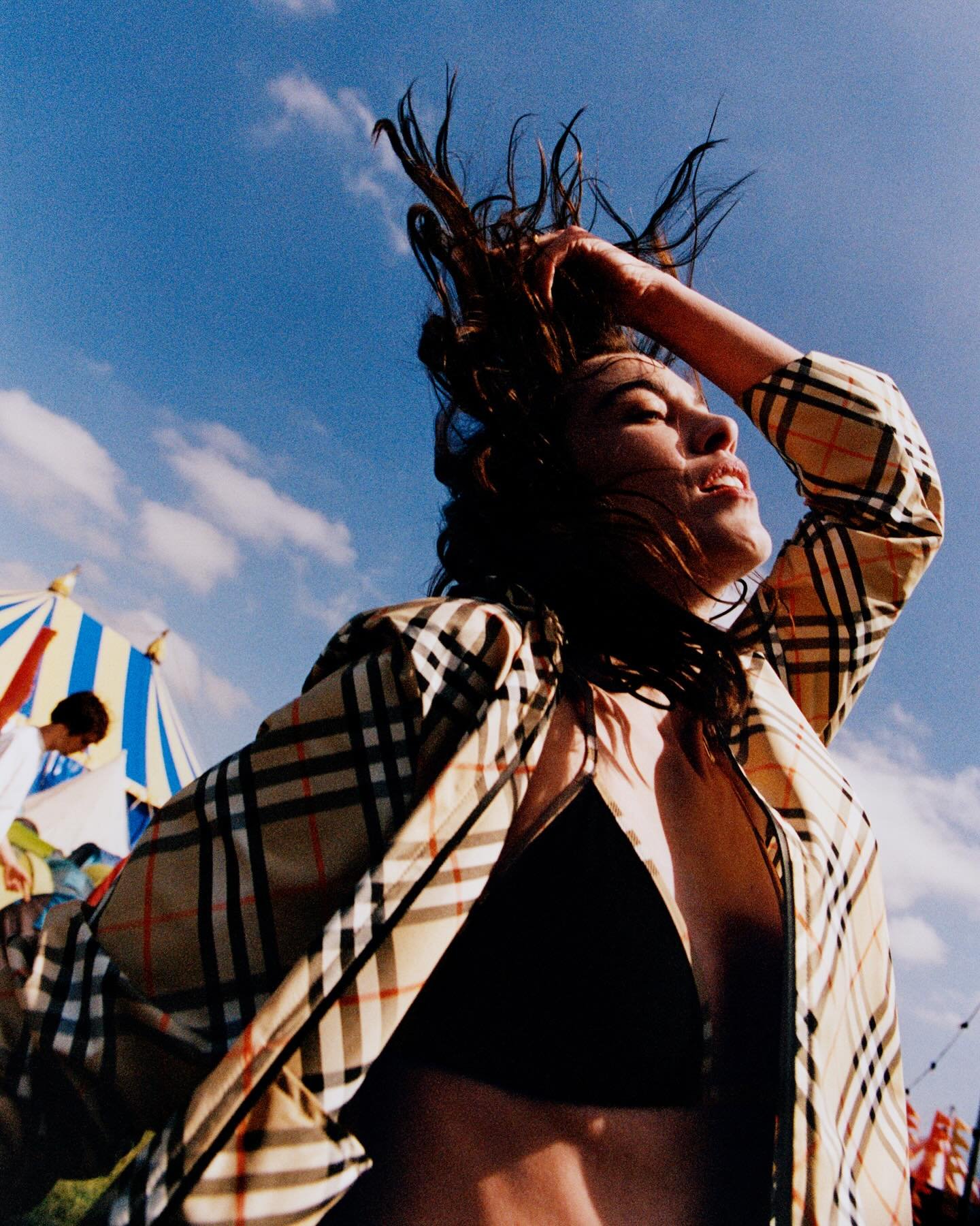 Festivais Burberry: Alexa Chung com roupa de padrão Burberry em um ambiente ensolarado com estrutura de festival ao fundo, sugerindo um evento ao ar livre.