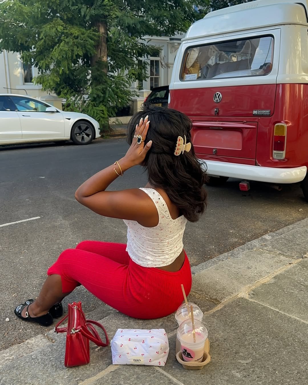 Mulher sentada na calçada com bolsa vermelha, roupas vibrantes e acessórios no cabelo. Kombi vintage ao fundo.