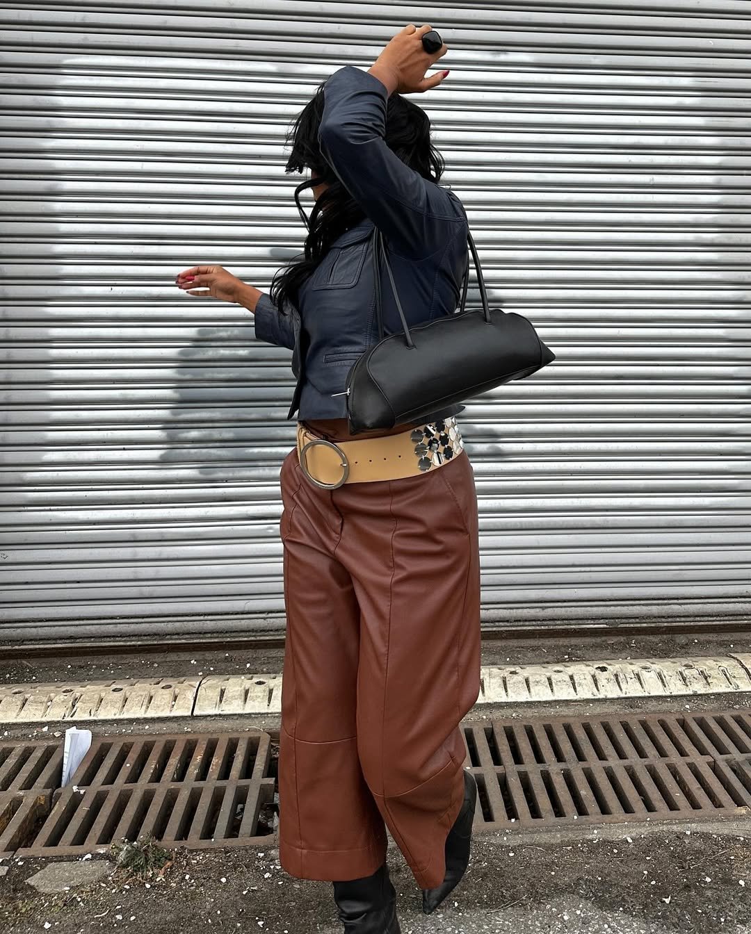 Pessoa usando casaco azul e calça marrom ajusta acessórios, incluindo bolsa preta e cinto com detalhe prateado. Fundo com porta de metal.