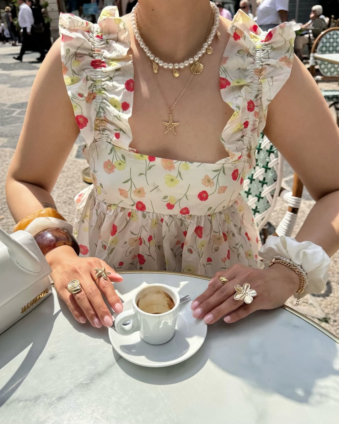 Vestido floral com acessórios, colares, anéis e pulseiras em destaque enquanto segura um café em mesa externa.