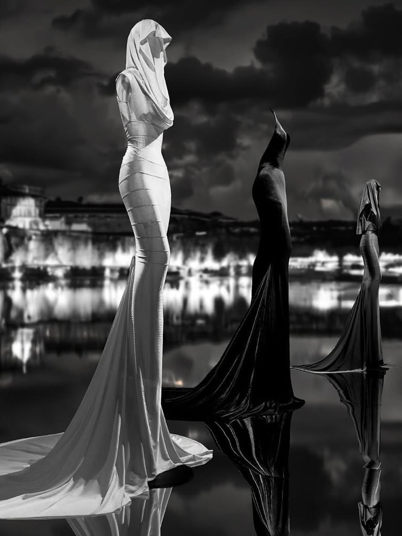 Vestidos elegantes em tecido drapeado, refletidos sobre uma água calma, com fundo urbano noturno. Alaïa