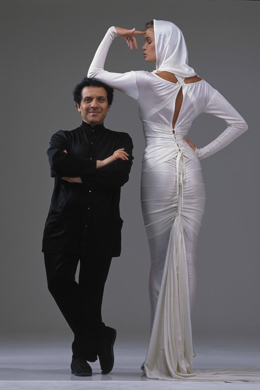 Estilista em pose confiante ao lado de modelo vestindo um elegante vestido branco Alaïa com capuz e costas abertas.