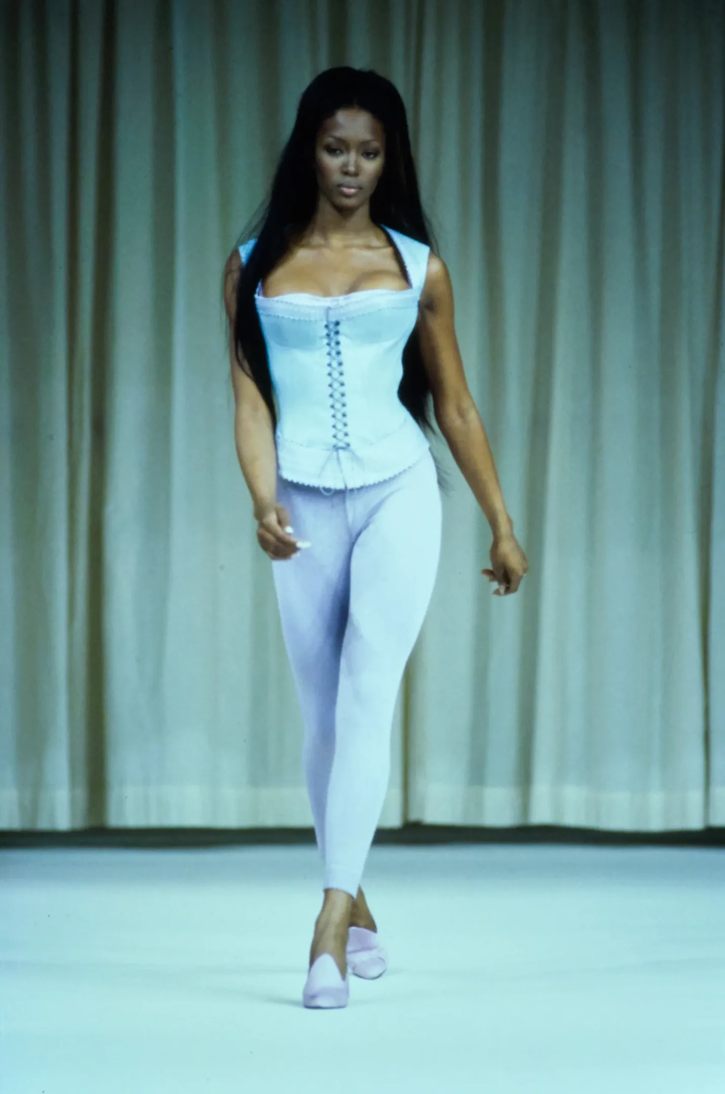 Modelo desfila com corset e leggings brancos da coleção Alaïa, fundo com cortinas neutras.