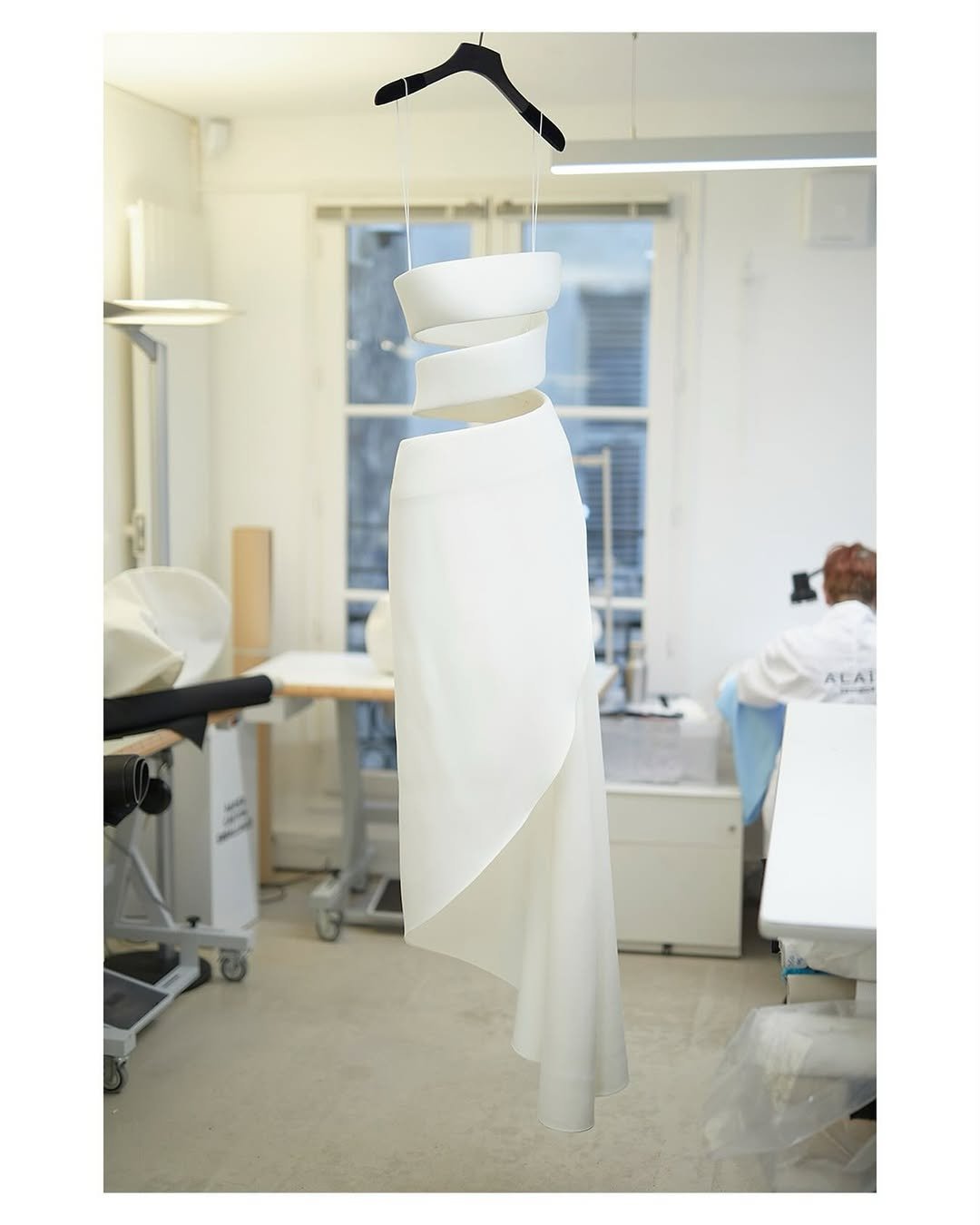 Vestido branco elegante Alaïa pendurado em cabide em ateliê, refletindo estilo sofisticado.