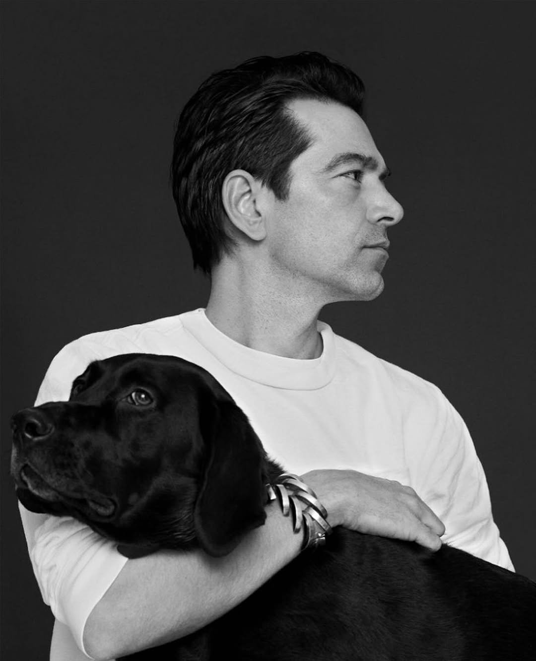 Homem em perfil abraça um cão preto, ambos em um cenário escuro. Ele usa uma pulseira estilo Alaïa.
