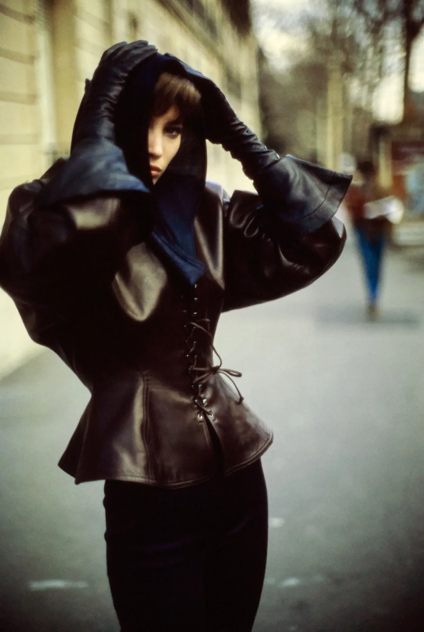 Pessoa usando um casaco de couro escuro com capuz e luvas, em uma rua urbana. Estilo remete à marca Alaïa.