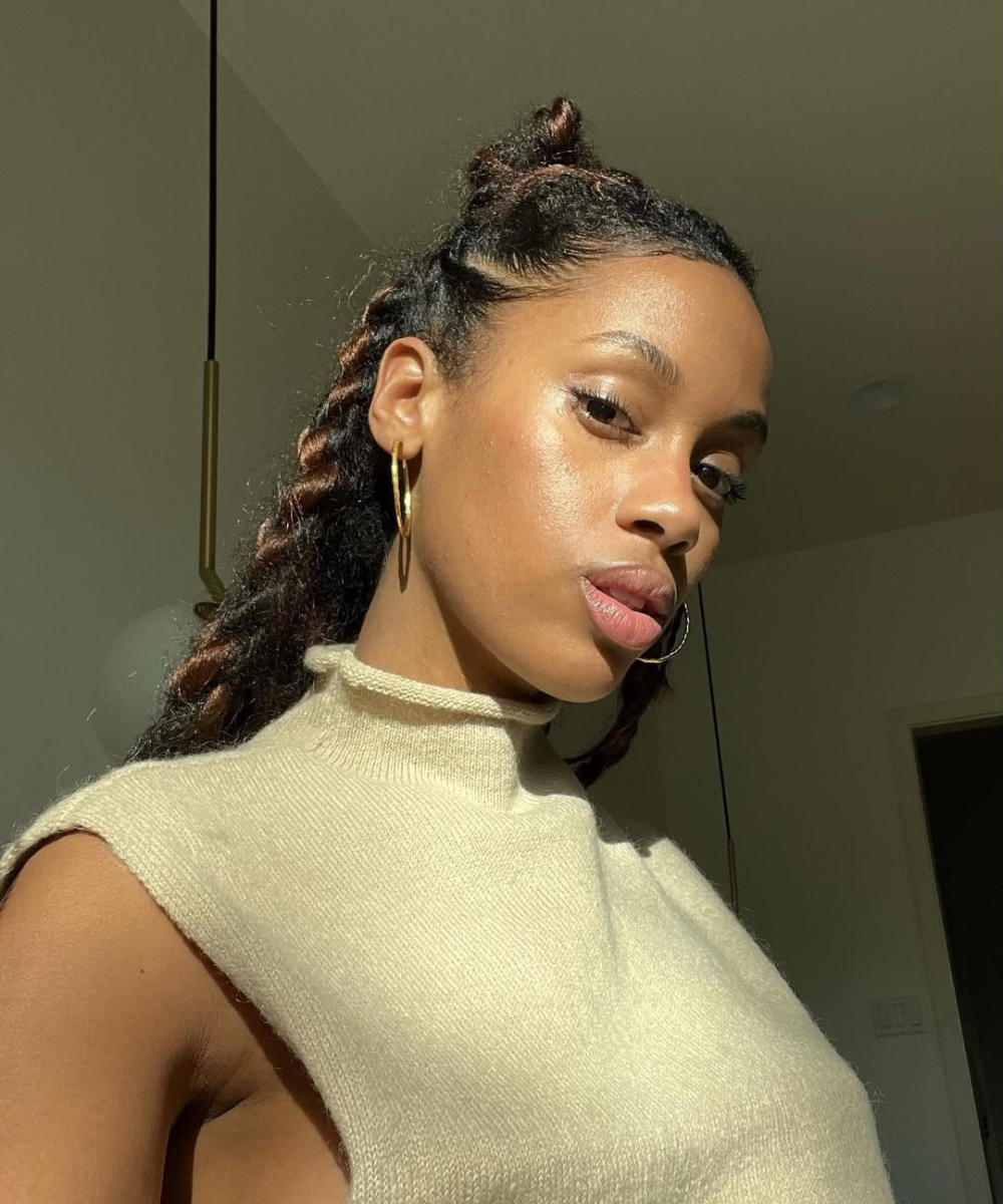 Mulher com cabelos trançados grandes, vestindo blusa de gola alta e brincos, sob luz natural. Alopecia por tração não visível.