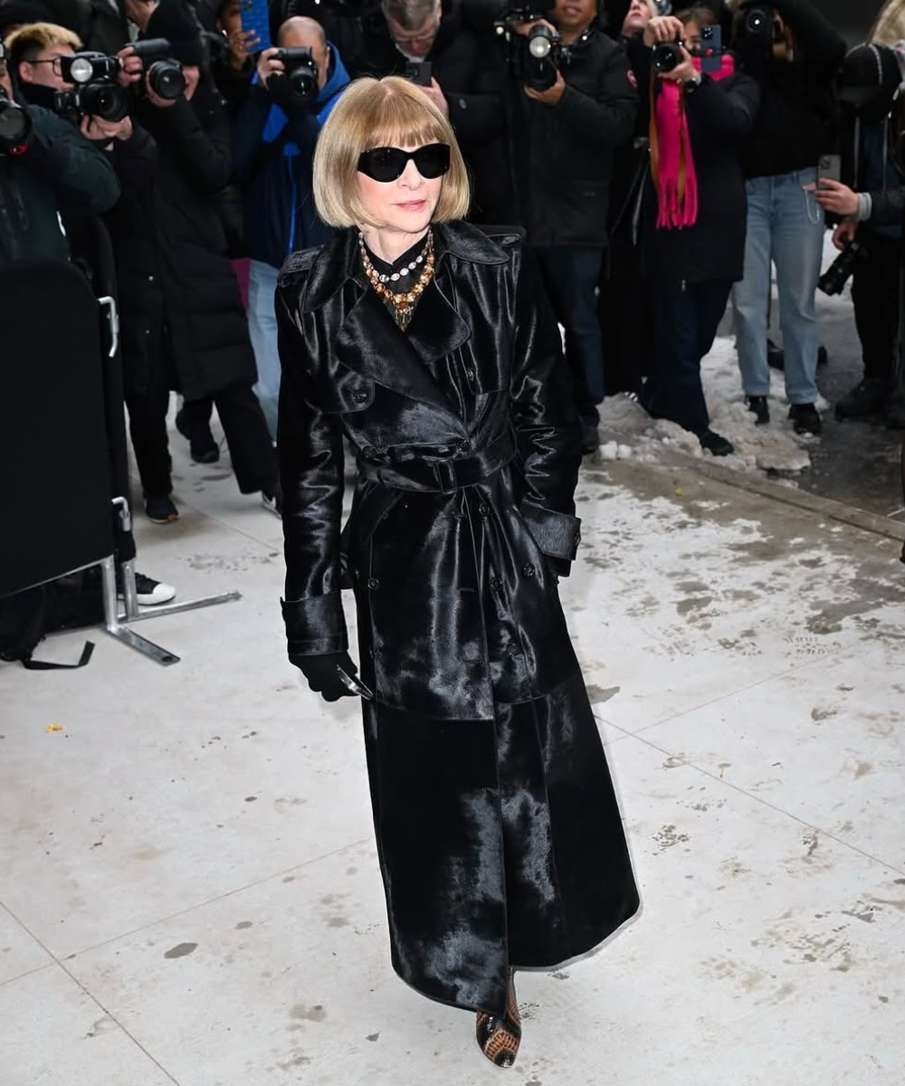 Anna Wintour usa casaco preto elegante e óculos escuros, cercada por fotógrafos.