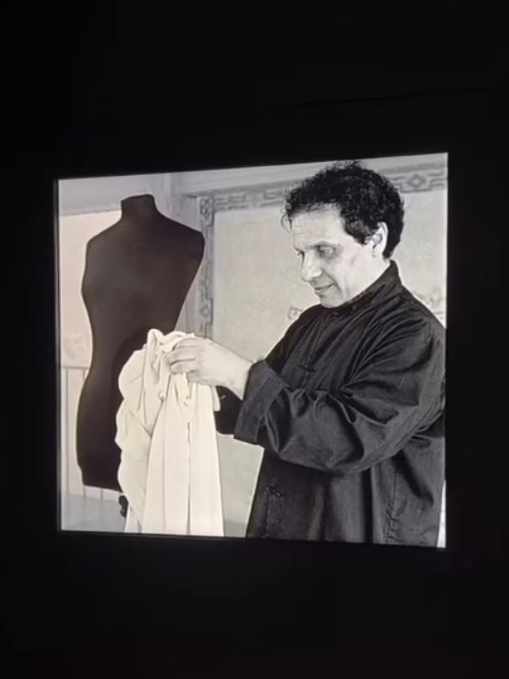 Um estilista segura uma peça de tecido branco próximo a um manequim, analisando o material. Alaïa