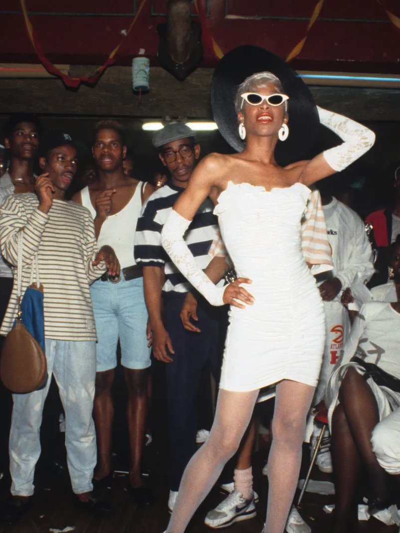 Pessoa posa com elegância em festa LGBTQIAPN+, usando vestido branco, óculos escuros e chapéu preto, rodeada por grupo animado.