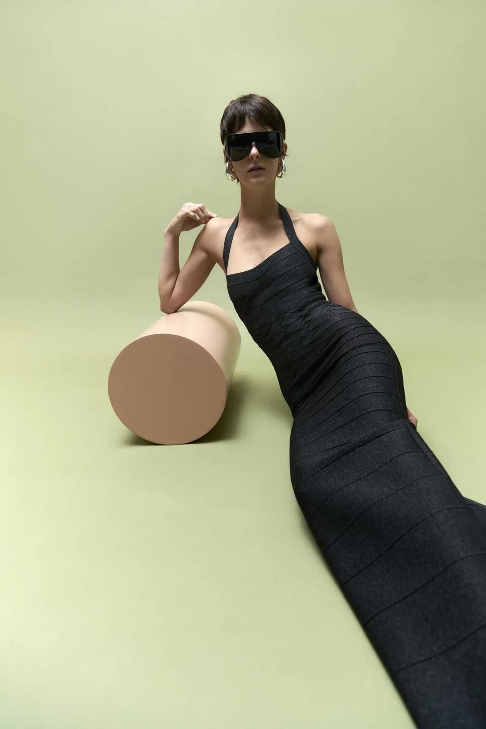 Pessoa em vestido preto ajustado e óculos de sol grandes, apoiada em cilindro de cor bege sobre fundo verde.