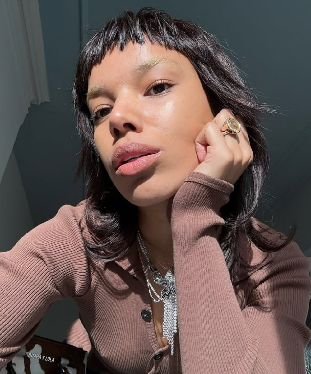 Uma pessoa com blusa marrom e colares posa sob luz natural, destacando uma pele impecável.