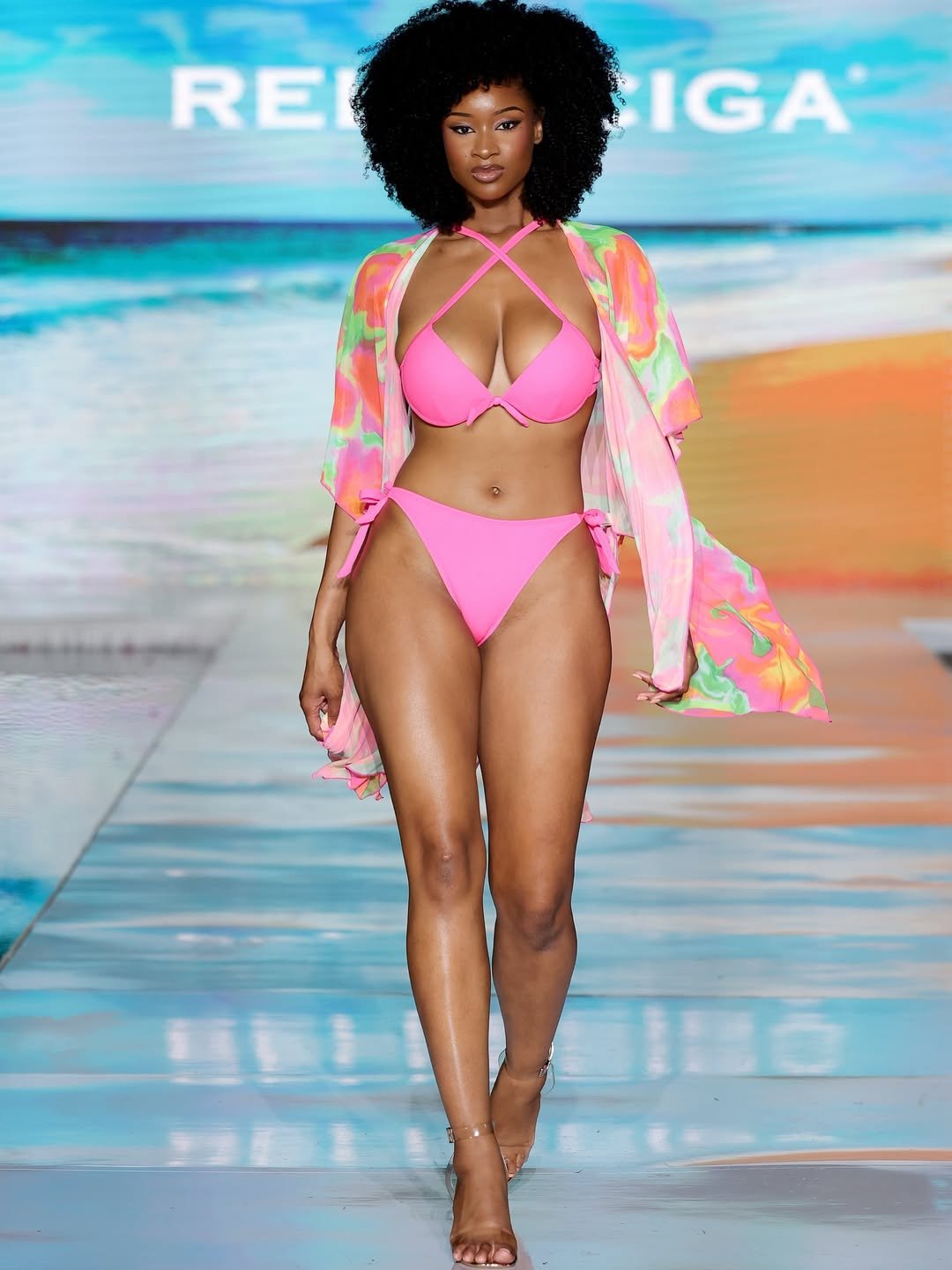 Modelo em passarela usando biquíni rosa brilhante e saída estampada, representando tendências de moda praia.