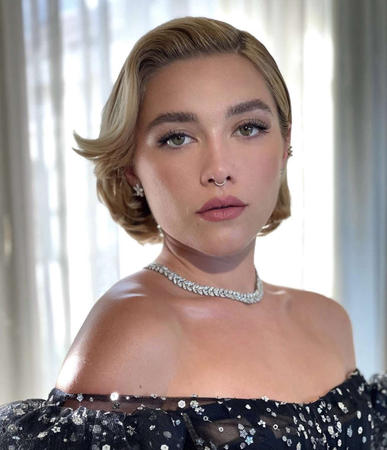 Mulher com cabelo bixie cut loiro, encaracolado nas pontas, usando colar brilhante e vestido preto com brilho.