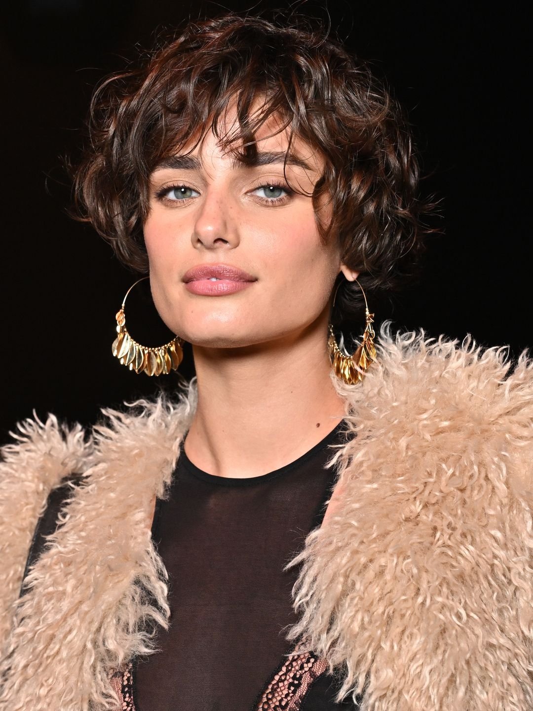 Pessoa com penteado bixie cut, usando brincos grandes e casaco de pele sintética.